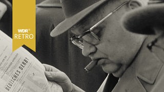 Eine Person mit einer Zigarre im Mund liest einen Zeitungsartikel mit der Überschrift "DEUTSCHES DERBY"