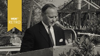 60er Jahre: Ein Mann steht hinter einem Podest und hält eine Ansprache.