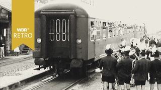 Ein Zug mit Fahrgästen, die aus den Fenstern winken. Neben dem Zug ein bayerisches Orchester.
