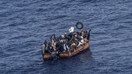 In einem kleinen, rostigen Boot sitzen viele Menschen, welche über das Mittelmeer versuchen, die europäische Küste zu erreichen.