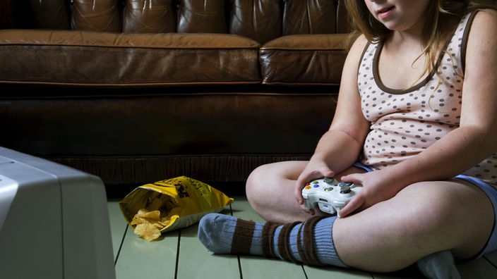 Ein übergewichtiges Kind sitzt vor einem Bildschirm und spielt ein Spiel. Daneben liegt eine offene Tüte Chips.