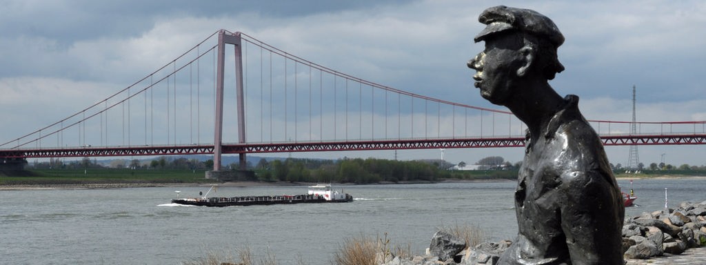 Die Bronzefigur Poortekeerl an der Emmericher Rheinpromenade, im Hintergrund die Hängebrücke
