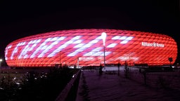 Zu sehen ist die Allianz Arena in der Nacht. Die rot-weiße Beleuchtung des Stadions ist zur Erinnerung an Franz Beckenbauer gewählt worden.