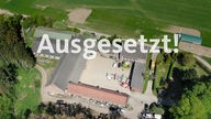 Schriftzug "Ausgesetzt!" vor einer Drohnenaufnahme eines Bauernhofs