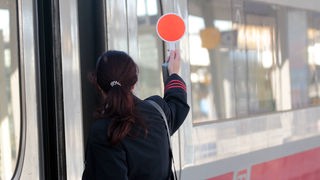 Eine Zugbegleiterin hält auf einem Bahnsteig die rote Kelle hoch