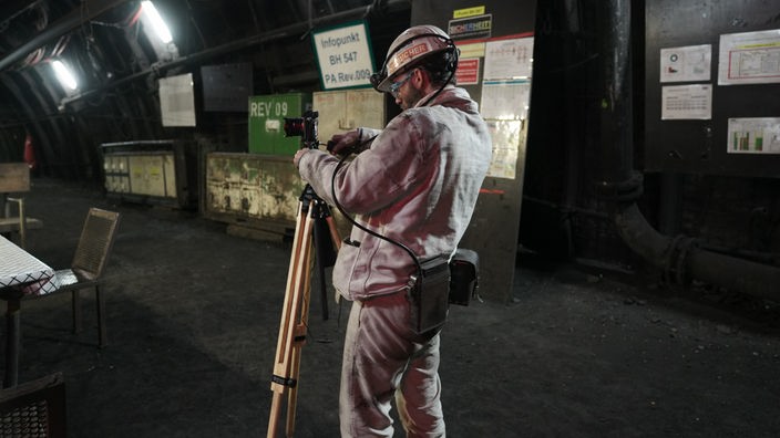 Ein Mann steht in einem weißen Arbeitsanzug, mit Schutzhelm und Kamera am Infopoint im Bergwerk