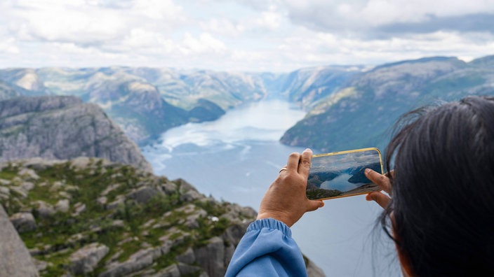 Frau steht auf dem Berg mit dem Felsen Preikestolen mit aussicht auf den Fjord Lysefjord und macht ein Foto