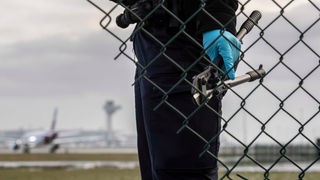 Ein Mann in Uniform mit Gummihandschuhen und einen Drahtschneider in der Hand steht an einem Zaun mit Loch darin. Im Hintergrund ein Flughafen.