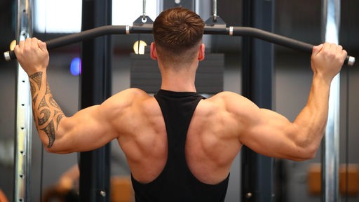 Ein junger Mann sitzt an einem Gerät im Fitnessstudio und trainiert seinen Rücken
