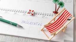 "Brückentag" geschrieben auf einem Kalender mit einem Liegestuhl und einer Palme