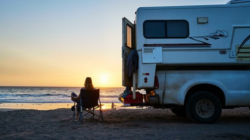 Eine Frau sitzt bei Sonnenuntergang neben einem Wohnmobil am Strand