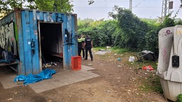 Müll liegt um einen alten Container herum, drei Mitarbeiter in Uniform begutachten das Umfeld