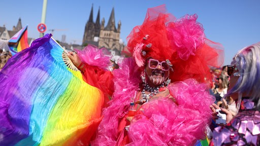 Bunt geschminkte und schrill gekleidete Teilnehmer*in bei der Parade des CSD in Köln, im Hintergrund der Dom