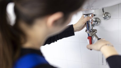 Eine Handwerkerin montiert den Abfluss eines Waschbeckens, sie hat eine Zange an das Rohr angesetzt.
