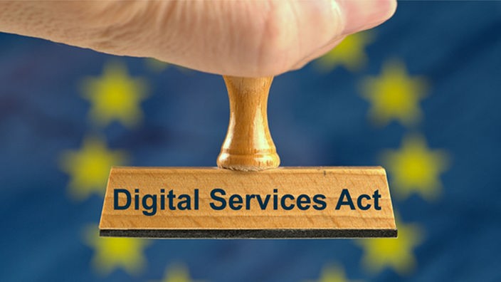 Ein symbolischer Holzstempel mit der Aufschrift "Digital Services Act", gehalten von einer Hand vor einer unscharfen EU-Flagge im Hintergrund. 