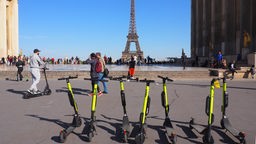 E-Scooter stehen auf einem Platz in Paris in einer Reihe, im Hintergrund ist der Eiffelturm zu sehen.