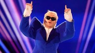 Ein Mann mit blond gefärbten Haaren, Sonnenbrille und blauem Anzug zeigt mit beiden Zeigefingern gen Himmel.