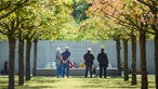 Menschen stehen am 25. Jahrestag in der Gedenkstätte vor der Gedenktafel der Opfer des ICE-Unglücks von Eschede unter Kirschbäumen. 