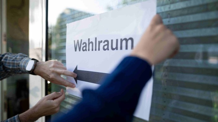 Wahlhelfer in NRW hängen einen Zettel, auf dem "Wahlraum" steht, auf.