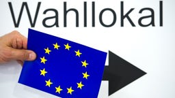 Eine Hand hält eine Karte mit der Europaflagge vor einem Plakat mit der Aufschrift "Wahllokal"