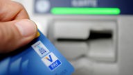 Eine Hand hält eine blaue Girokarte in Richtung eines Bankautomaten.