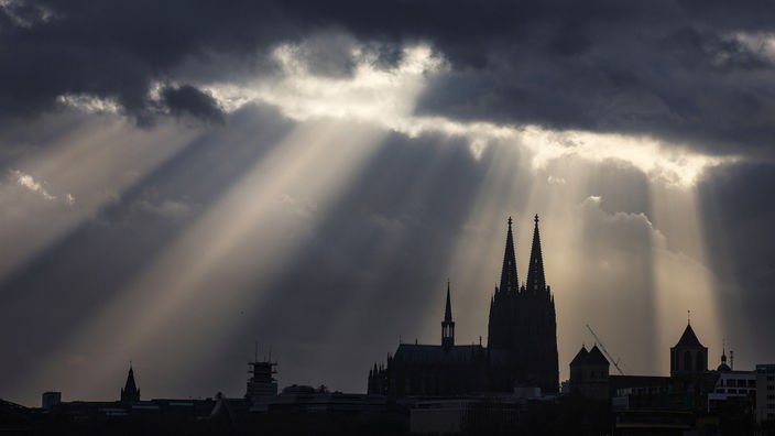 Sonnenstrahlen durchbrechen eine Wolkenschicht. Das Stadtbild Kölns zeigt sich als Silhouette. 