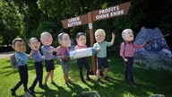 Aktivisten der Organisation Oxfam tragen Masken der G7-Spitzenpolitiker und stehen in Wanderkleidung vor einem Wegweiser, der in der einen Richtung Leben retten anzeigt, in entgegengesetzter Richtung Profite ohne Ende anzeigt