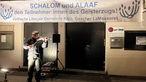 Ein Mann spielt Geige. Hinter ihm hängt ein Banner mit dem Aufdruck "Schalom und Alaaf den Teilnehmer:innen des Geisterzugs! Jüdische Liberale Gemeinde Köln, Gescher LaMassoret"