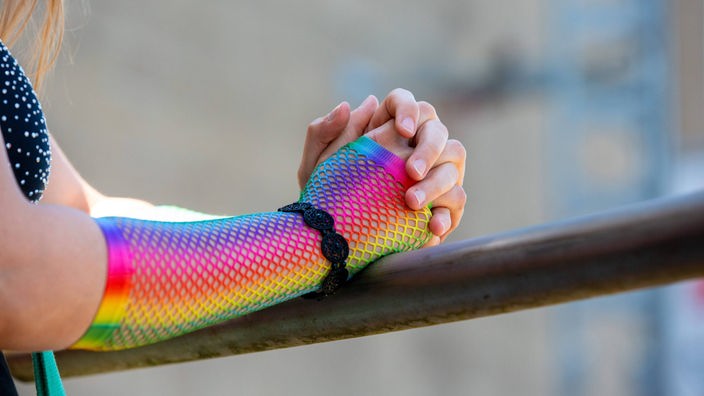 Frau mit regenbogenfarbener Unterarmbedeckung lehnt an einem Geländer.
