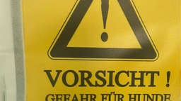 Gelbes Schild mit Warnzeichen und der Aufschrift "Vorsicht! Gefahr für Hunde!"