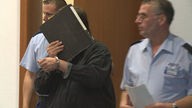 Im Gerichtssaal: Der Angeklagte befindet sich in der Mitte des Bildes und schützt sein Gesicht mit einem Ordner, er hat Handschellen um und wird von zwei Polizisten in den Saal eskortiert.