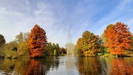 Die Bäume im Stadtpark Gütersloh tragen Blätter im Herbst-Farbenkleid. Im Wasser davor schießt eine Fontäne empor