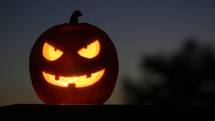 Als Halloween-Dekoration steht ein ausgehöhlter, beleuchteter Kürbis mit einem geschnitzten grinsenden Gesicht in einem Garten