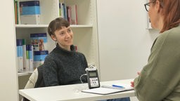 Hebamme Klara sitzt vor einem Bücherregal mit Fachliteratur zur Geburtshilfe. Auf dem Tisch vor ihr steht ein Aufnahmegerät, ihr gegenüber sitzt die Reporterin.