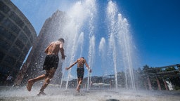 Zwei Jungs in schwarzen Hosen laufen oberkörperfrei durch die Fontäne einer städtischen Springbrunnenanlage.