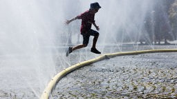 Ein Junge springt über einen Feuerwehrschlauch aus dem Wasser raus spitzt um sich gegen die Hitze abzukühlen.