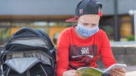 Junge mit Maske und Käppi sitzt auf Schulhof und liest in einem Buch.