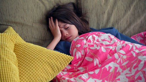 Ein krankes junges Mädchen liegt eingedeckt auf dem Sofa und hält sich die Hand an den Kopf