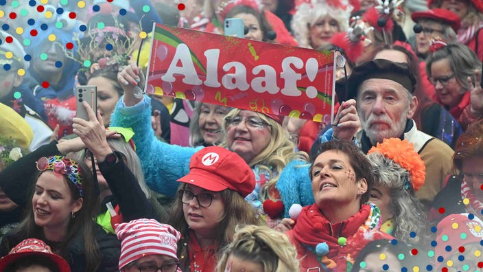 Verkleidete Menschen feiern die Sessionseröffnung am 11.11.23 in Köln. Eine Frau hält ein kleines Banner mit der Aufschrift "Alaaf!" hoch