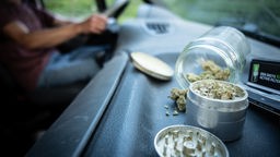 Ein offenes Glas mit Cannabisblüten und ein Grinder liegen auf dem Armaturenbrett eines Autos.