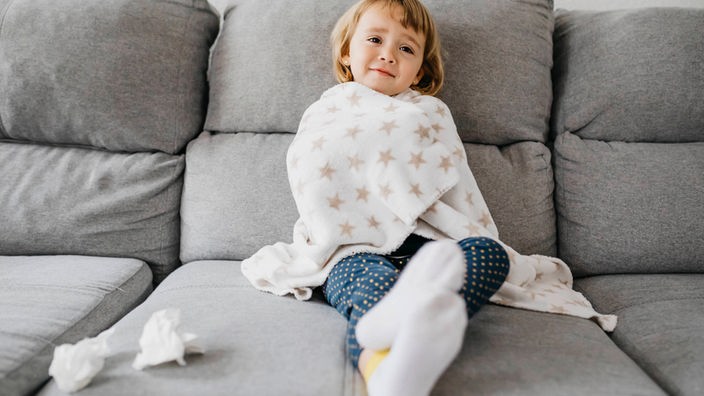 Ein krankes Kind sitzt in eine Decke gewickelt auf dem Sofa, Taschentücher neben legen ihm 
