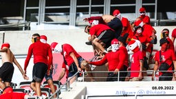 Eine Gruppe maskierter Köln-Fans klettet über Absperrungen und Sitze. Unten rechts das Datum "08.09.2022"