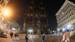 Der Kölner Dom bei Nacht ohne Beleuchtung. Im Vordergrund sind vorbei laufende Menschen zu sehen.