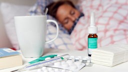 Medikamente und ein Fieberthermometer liegen auf einem Nachttisch, eine Frau liegt dahinter im Bett.