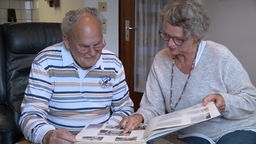 Ein älterer mann und eine Frau sitzen am Fliesentisch und schauen sich ein Fotoalbum an, was vor ihnen liegt.