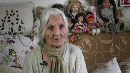 Theresia Neger, eine weißhaarige, ältere Frau, sitzt auf einem Sofa, im Hintergund sind Puppen zu sehen