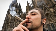 Ein Mann steht vor dem Kölner Dom und raucht einen Joint