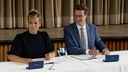 Mona Neubaur und Hendrik Wüst unterzeichnen den Koalitionsvertrag an einem Tisch mit weißer Tischdecke sitzend