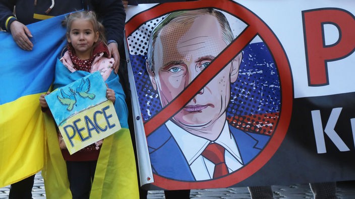 Szene einer Demonstration in Münster: Ein kleines Mädchen trägt ein Schild in blau undgelb, auf dem zwei Friedenstauben und PEACE abgebildet sind, daneben ist ein Anti-Putin-Transparent zu sehen. 