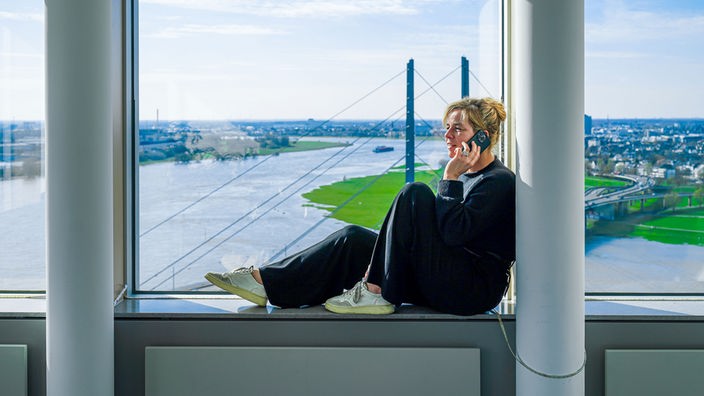 Gestelltes Foto: Mona Neubaur sitzt in ihrem Ministerium auf der Fensterbank und telefoniert mit dem Smartphone, im Hintergrund ist der Rhein zu sehen. 